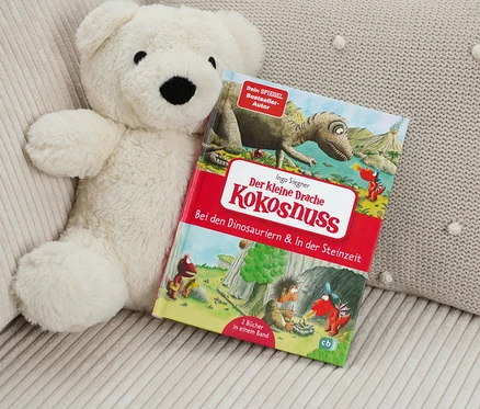 Un ours en peluche est posé à côté du livre «	Der kleine Drache Kokosnuss – Bei den Dinosauriern & In der Steinzeit	» (Le petit dragon Noix de coco – Chez les dinosaures et à l’âge de pierre, en allemand).
