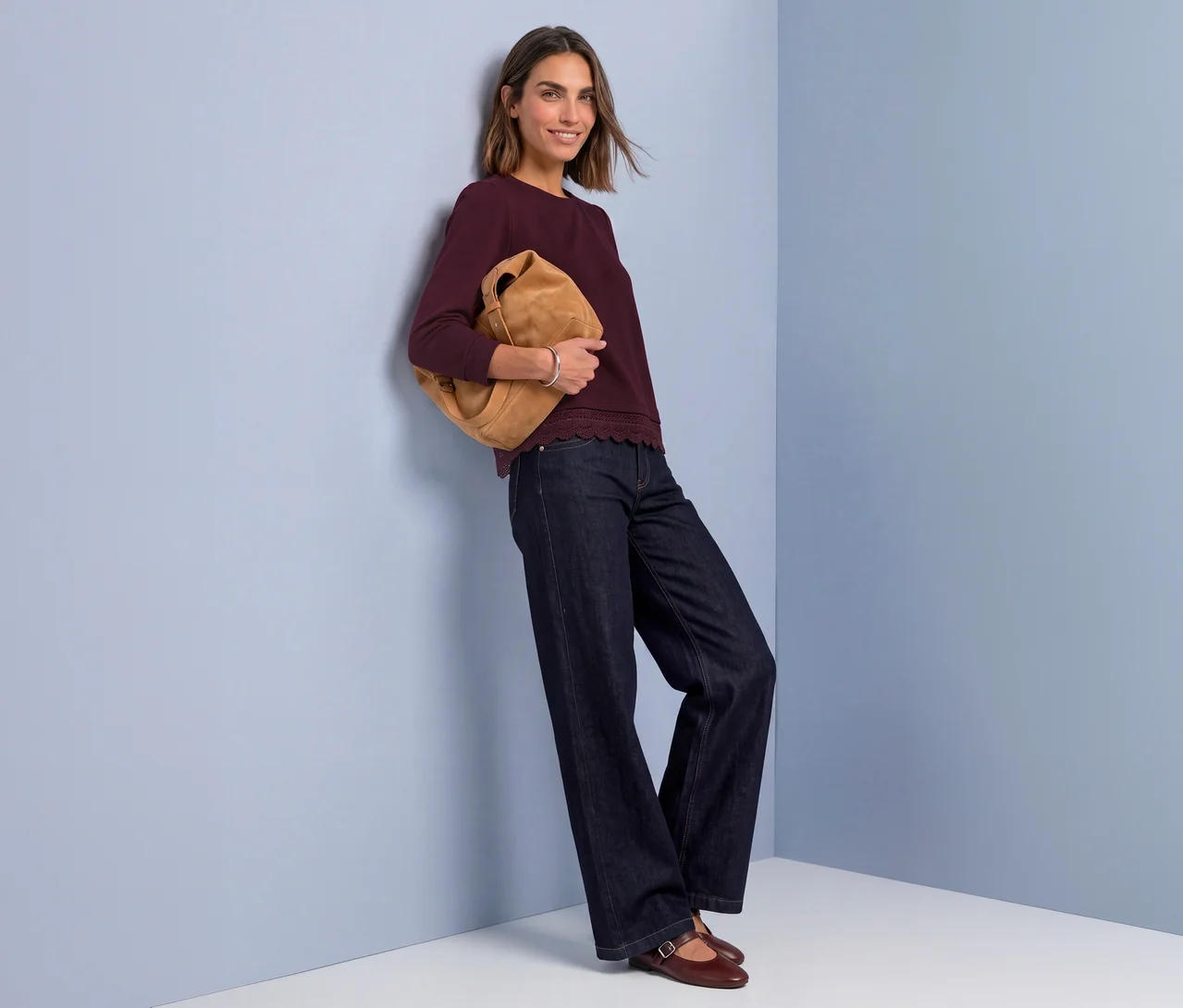 Femme souriante en t-shirt jersey 3/4 Street One rouge, jean large, tenant un sac marron, appuyée contre un mur bleu.