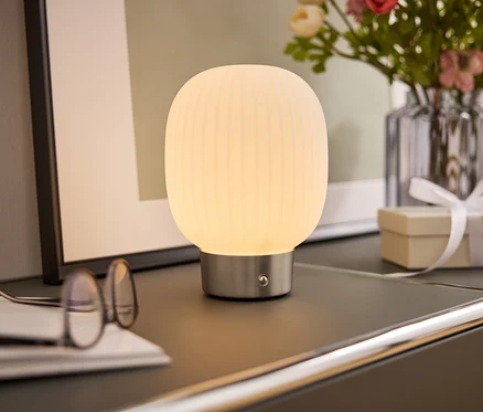 Une lampe de chevet avec un abat-jour blanc est posée sur une armoire à côté de lunettes, d'un livre, d'un vase de fleurs et d'une boîte cadeau.