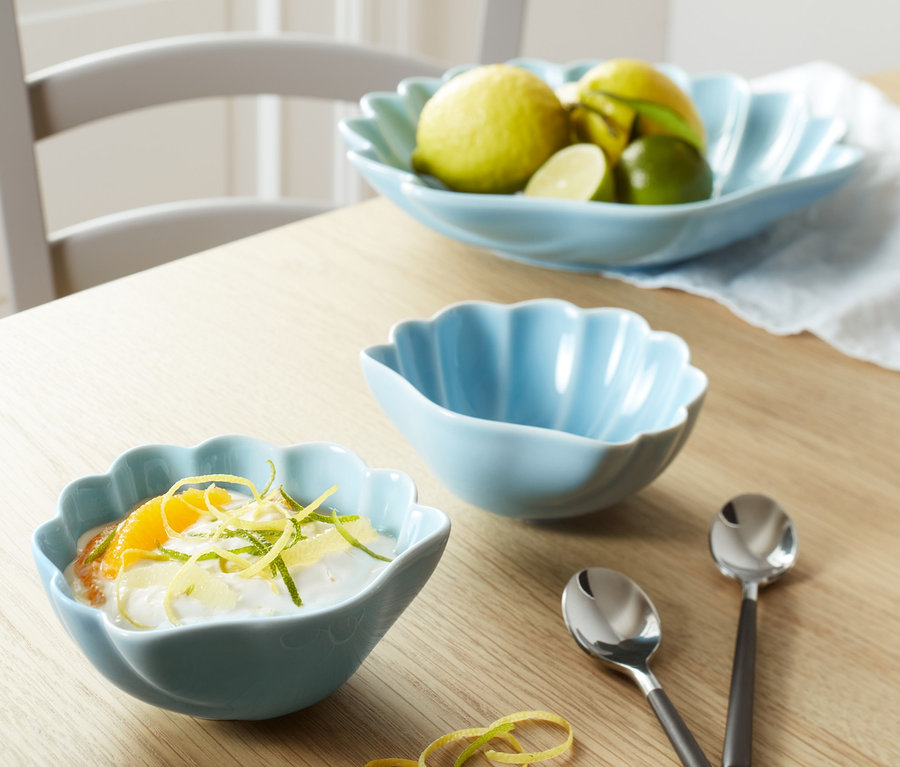 Sur une table, deux coupelles bleues et des cuillères. Une coupelle est remplie de dessert, l'autre est vide. À l'arrière-plan, une plus grande coupelle contient des fruits.