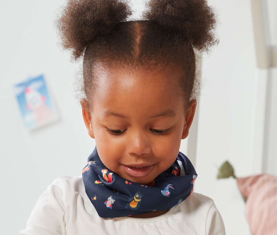 Fillette assise par terre jouant avec des cubes en bois. Elle porte un foulard multifonction avec imprimé intégral.