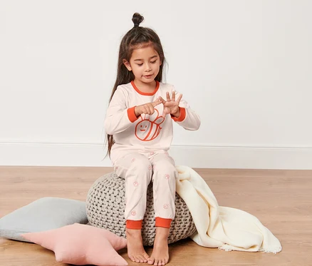 Une fillette assise sur un pouf compte sur ses doigts. Elle porte un pyjama rose et bleu pour tout-petits.