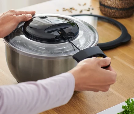 Une personne utilise une essoreuse à salade en inox avec couvercle dans une cuisine.