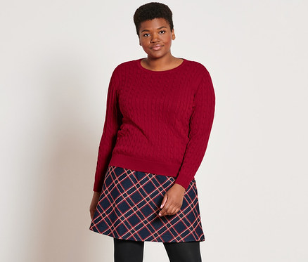 Une femme est debout et porte un pull en maille torsadée rouge, une jupe en jacquard à carreaux, 2 paires de collants opaques noirs et des bottines en cuir.