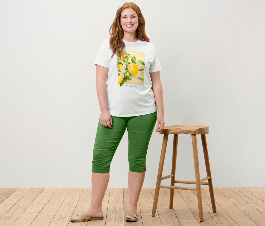 Une femme pose à côté d'un tabouret en bois, portant un t-shirt à imprimé et un pantalon 3/4 stretch vert.