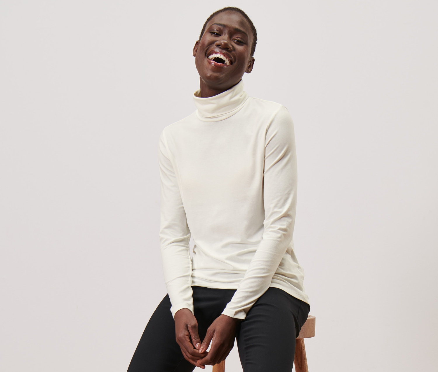 Femme assise sur un tabouret portant un t-shirt col roulé crème, un pantalon noir et des mocassins noirs.