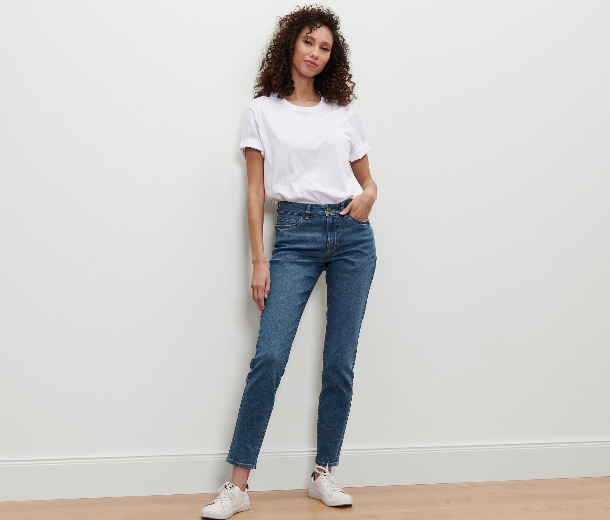 Une femme appuyée contre un mur porte un jean slim en denim « Fit Emma », denim bleu moyen, un t-shirt blanc et des baskets.
