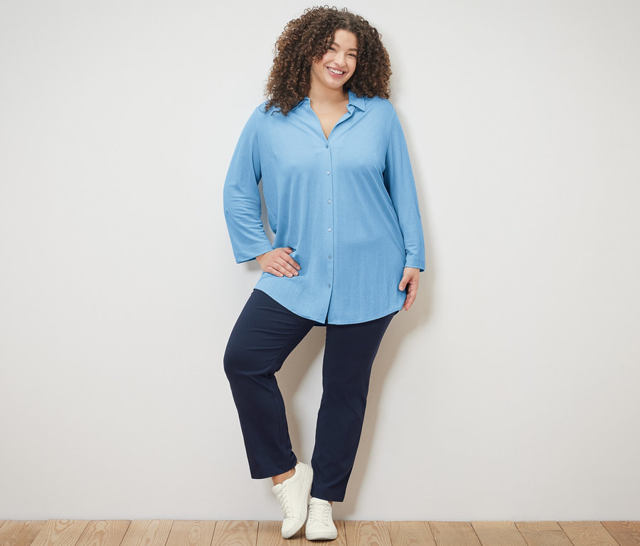 Femme posant sur un plancher en bois portant un chemisier en jersey piqué bleu clair et un pantalon stretch bleu marine.