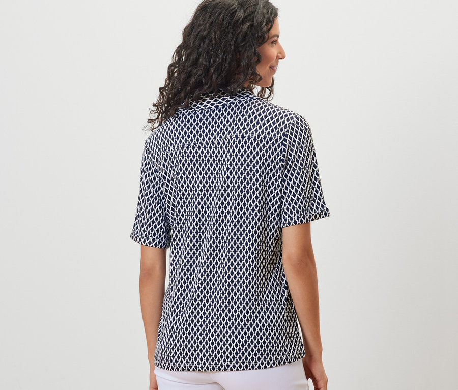 Vue arrière d'une femme aux cheveux ondulés foncés portant un chemisier imprimé avec patte de boutonnage et un pantalon blanc.