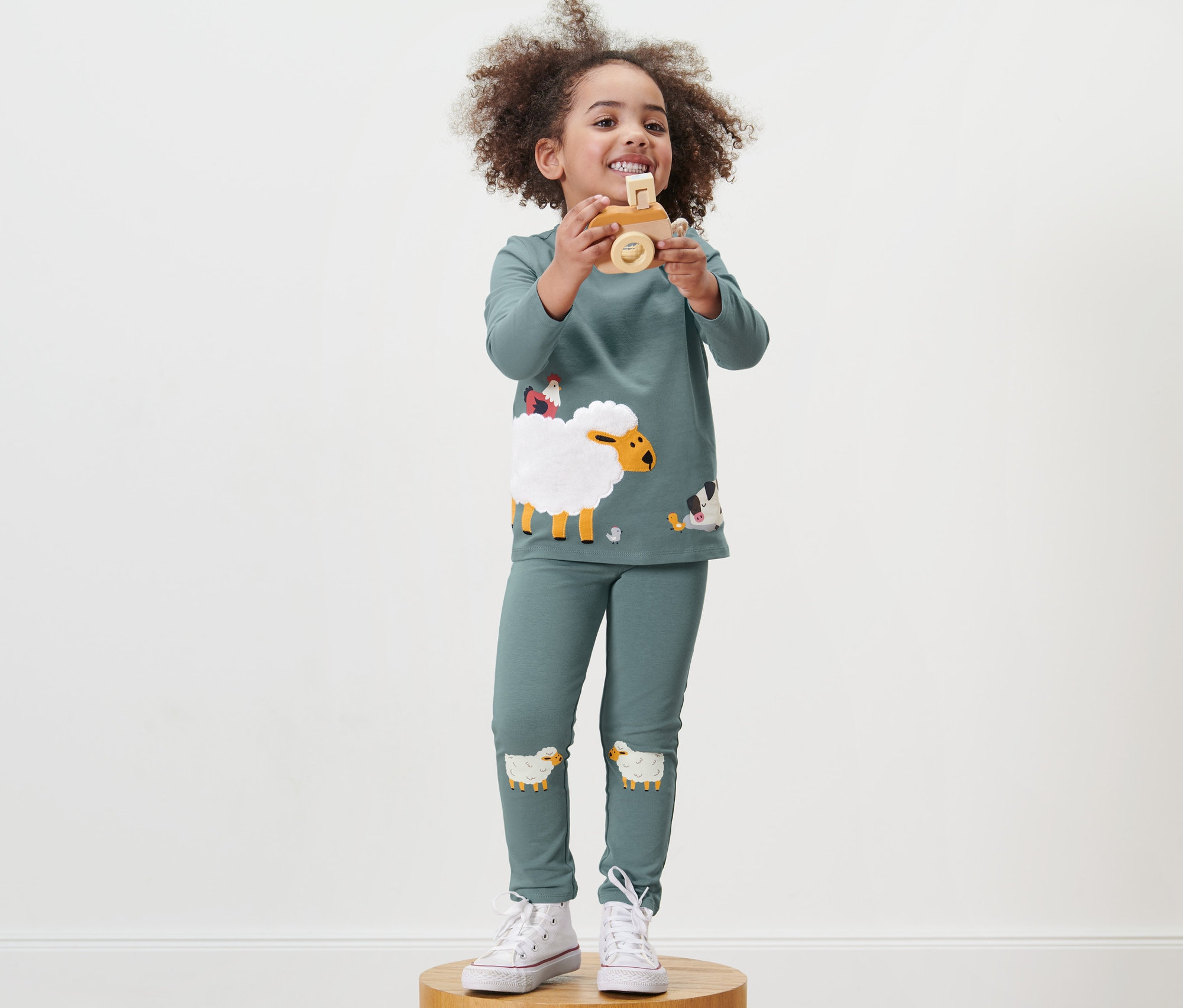 Une fille se tient sur un socle en bois et tient un jouet en bois. Elle porte un tregging pour tout-petits.