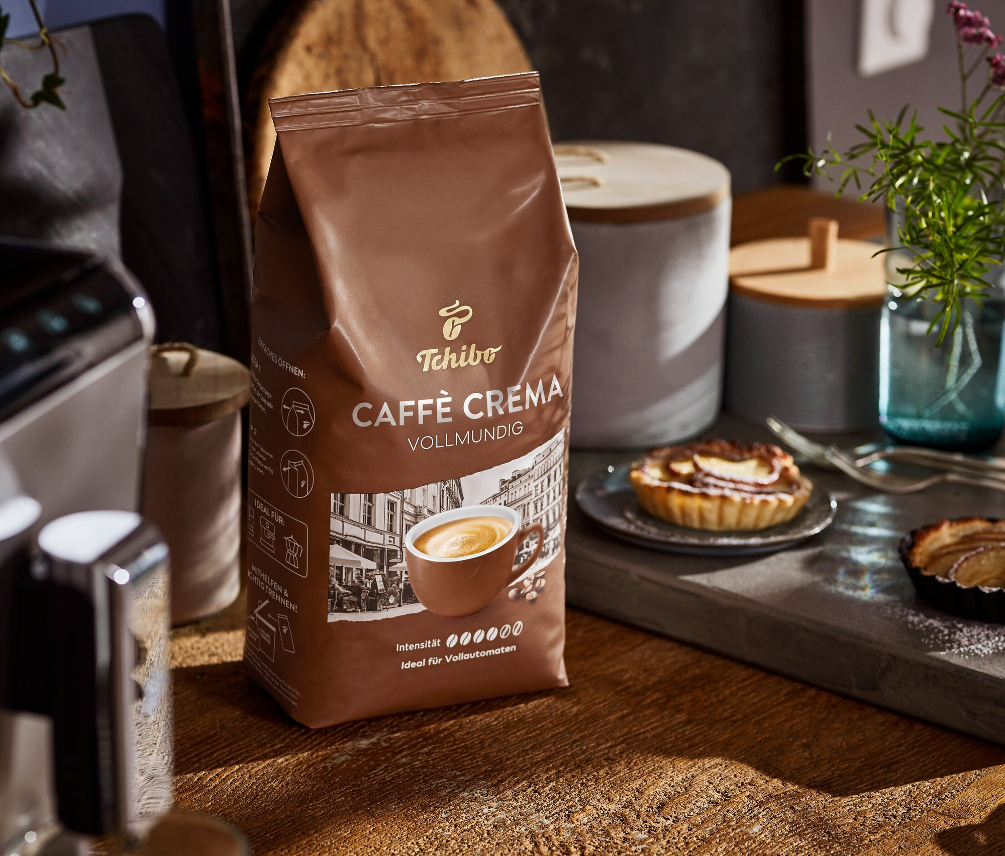 Un paquet de Caffè Crema Vollmundig est posé sur une table en bois à côté d'une machine à café et d'assiettes contenant des gâteaux.