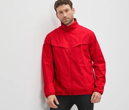 Un homme appuyé contre un mur portant une veste technique coupe-vent rouge.