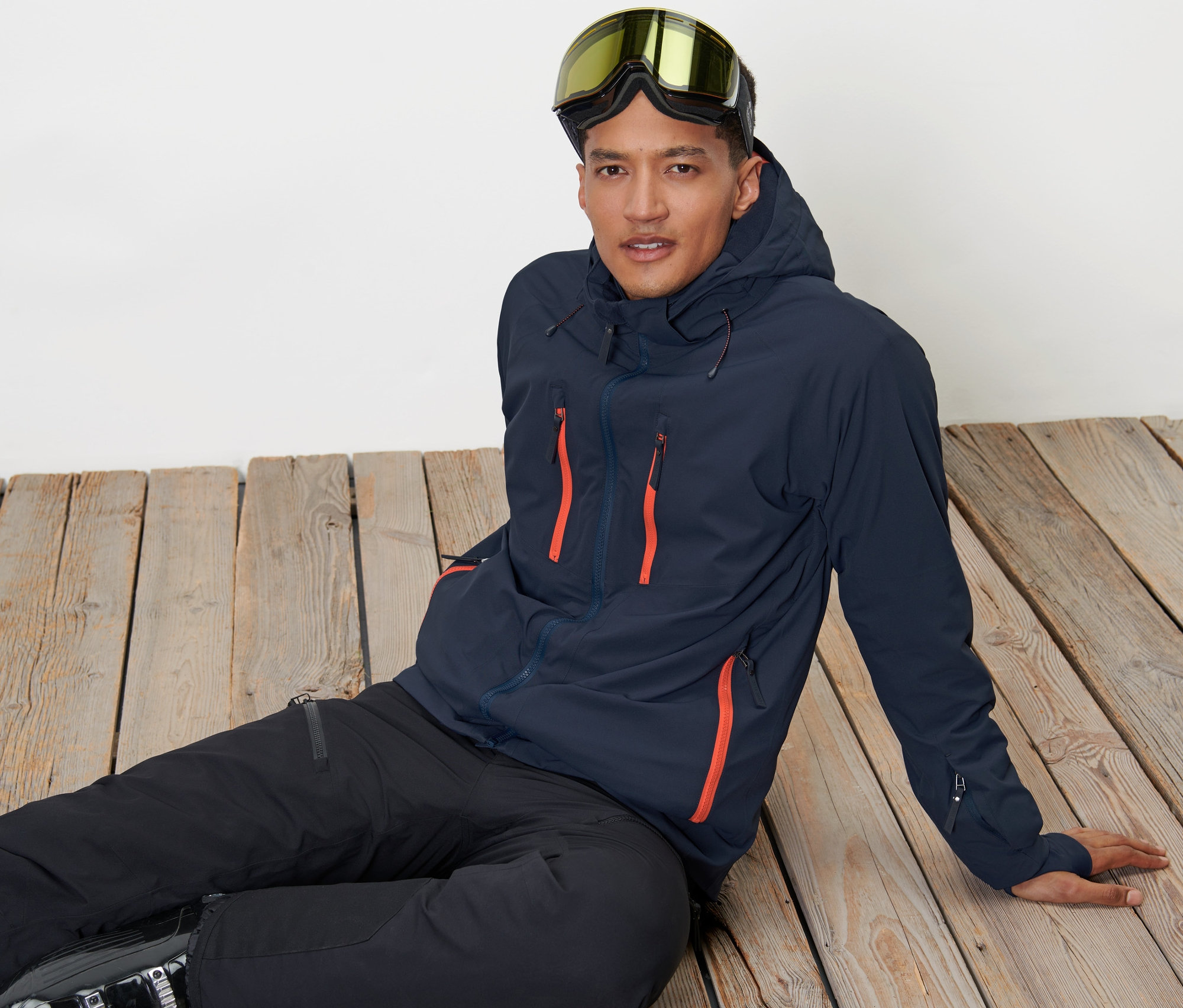 Un homme est assis sur un plancher en bois, portant une veste de ski high-tech et des lunettes de ski.