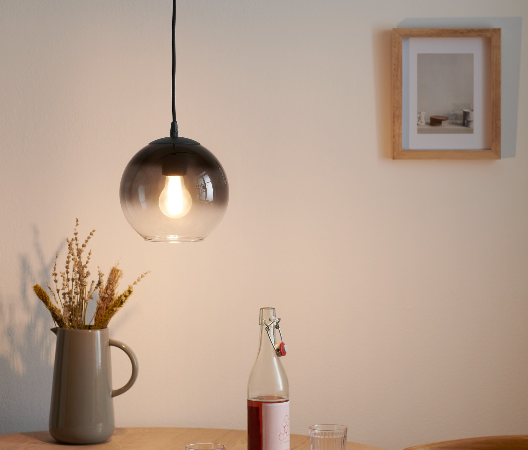 Une lampe à suspension avec abat-jour en verre fumé est suspendue au-dessus d'une table en bois avec un vase de fleurs, une bouteille et deux verres.