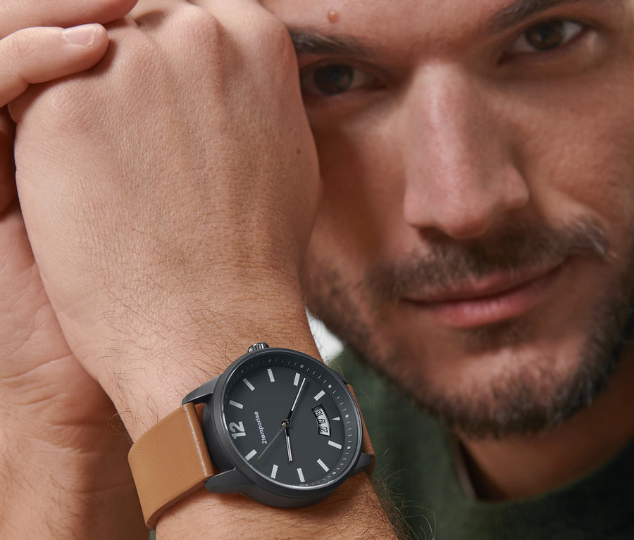 Un homme regarde la caméra et porte une montre style pilote noire avec un bracelet en cuir marron au poignet.