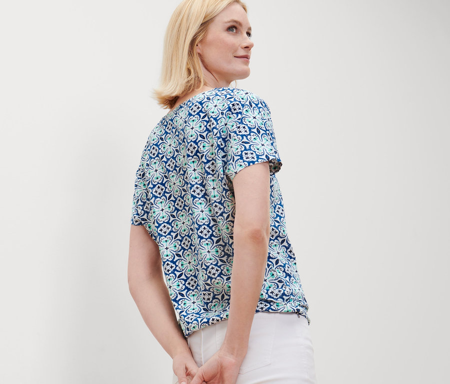 Une femme blonde pose en blouse avec un imprimé intégral et un pantalon blanc.