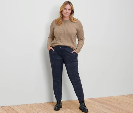 Une femme pose en pull en maille torsadée, crème chiné, pantalon de jogging et bottines en cuir.