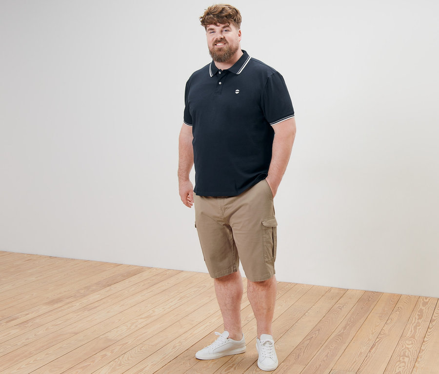 Un homme debout, une main dans la poche, portant un polo en piqué bleu marine et un short cargo beige.