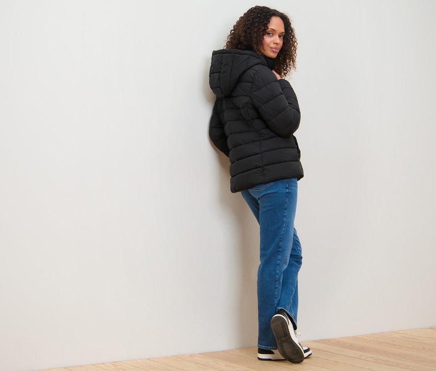 Femme appuyée contre un mur portant une veste matelassée à capuche.