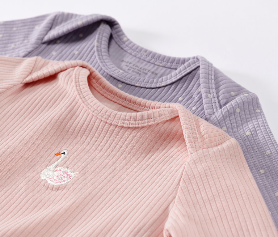 Gros plan sur deux t-shirts à manches longues pour bébé, rose et violet. Le t-shirt rose a un cygne brodé.