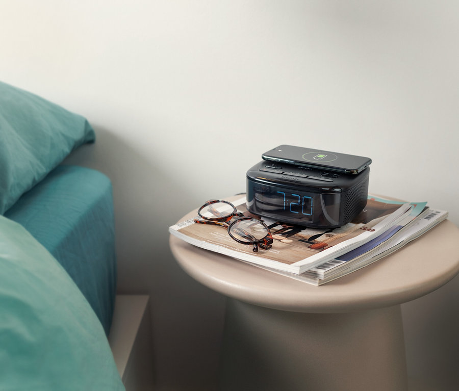 Sur une table de chevet se trouvent un radio-réveil DAB Philips « TAR7705 » avec un téléphone dessus et des lunettes à côté.