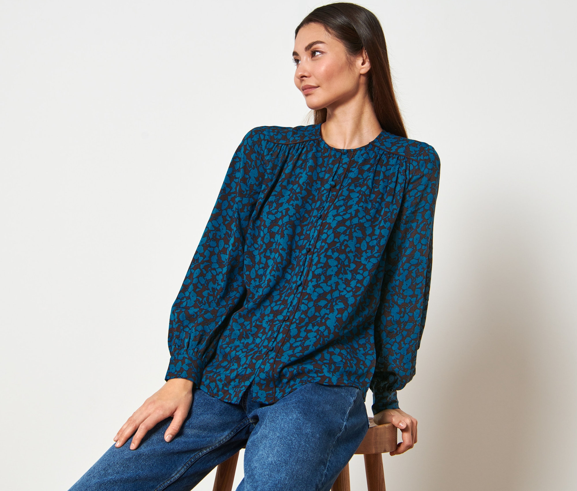 Une femme est assise sur un tabouret et porte une blouse à imprimé intégral et un jean.