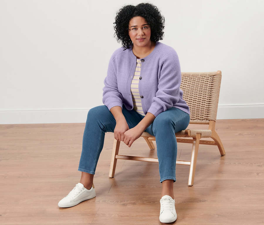 Femme assise sur une chaise en osier portant un t-shirt rayé beige et lilas à manches 3/4, un cardigan en maille, un jegging et des baskets en cuir.