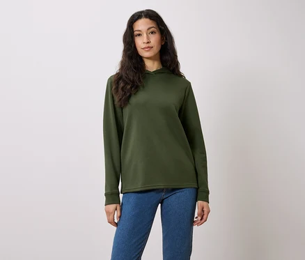 Une femme aux longs cheveux bruns pose dans un sweat à capuche vert olive et un jean bleu sur un fond blanc.