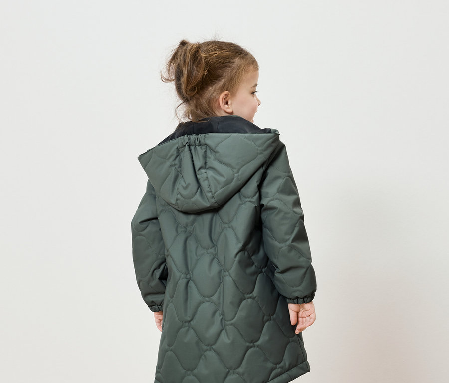 Vue de dos, une petite fille aux cheveux attachés en queue de cheval porte une veste matelassée verte à capuche.