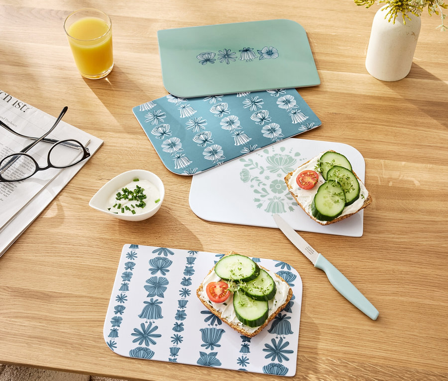Quatre planches à petit-déjeuner sont posées sur une table en bois, dont deux avec des tartines. On aperçoit également un verre de jus d'orange, un bol de sauce et un couteau.