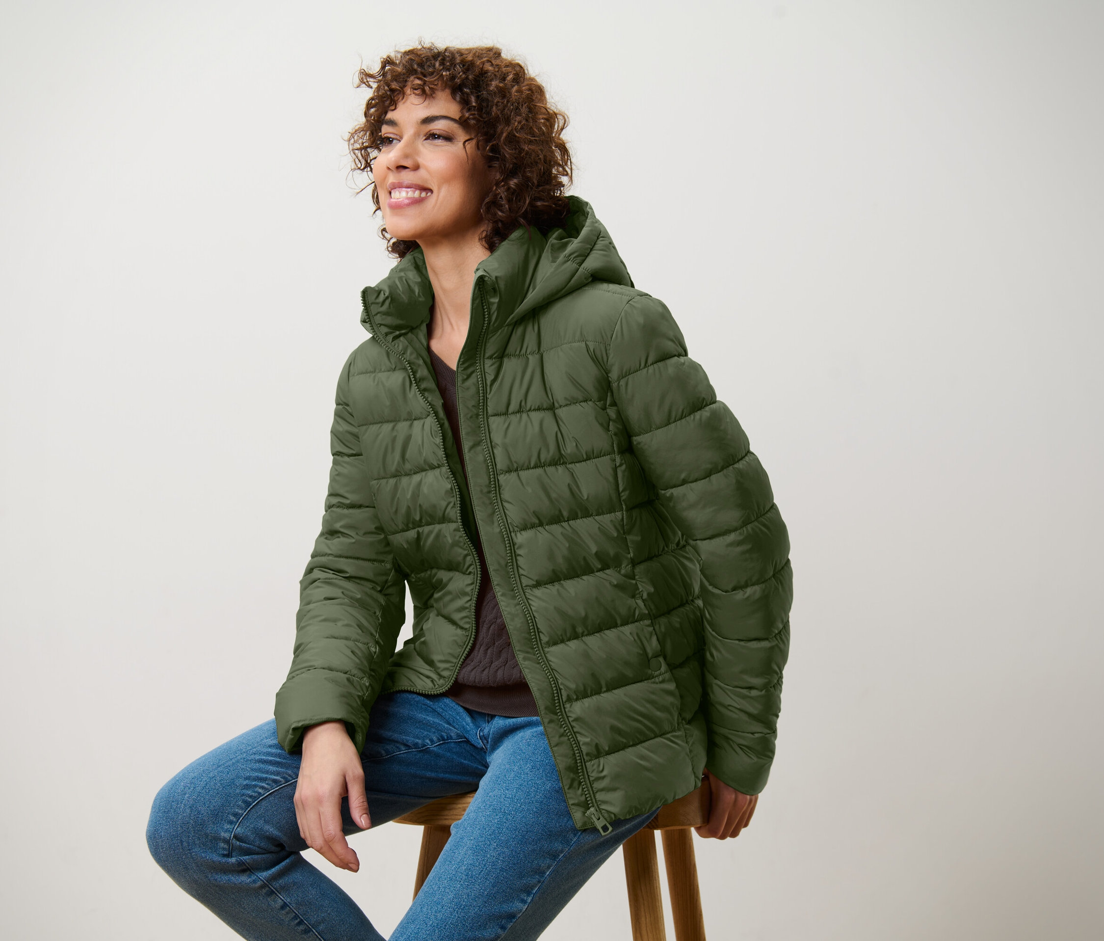 Une femme souriante est assise sur un tabouret et porte une veste matelassée vert olive et un jean.