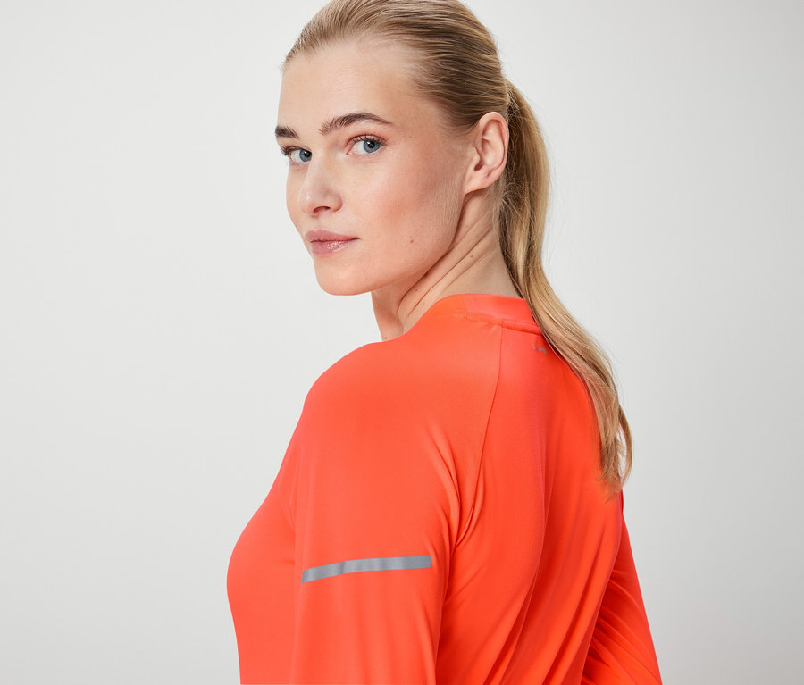 Une femme blonde regarde par-dessus son épaule et porte un haut orange à manches longues.