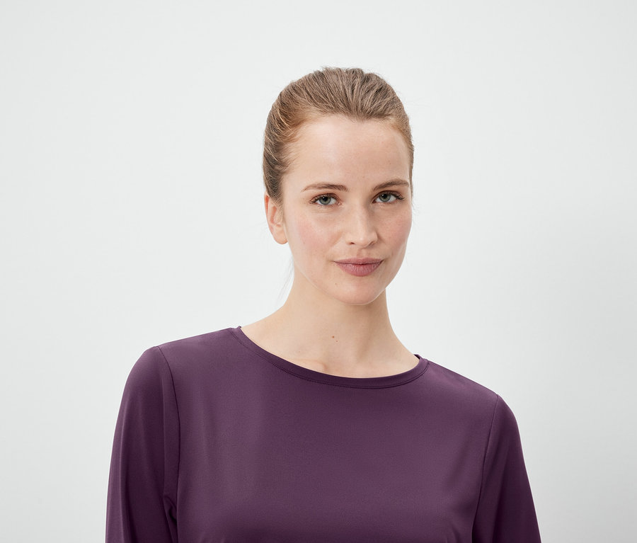Portrait d'une femme aux cheveux tirés en queue de cheval, portant un haut violet à manches longues sur un fond blanc.