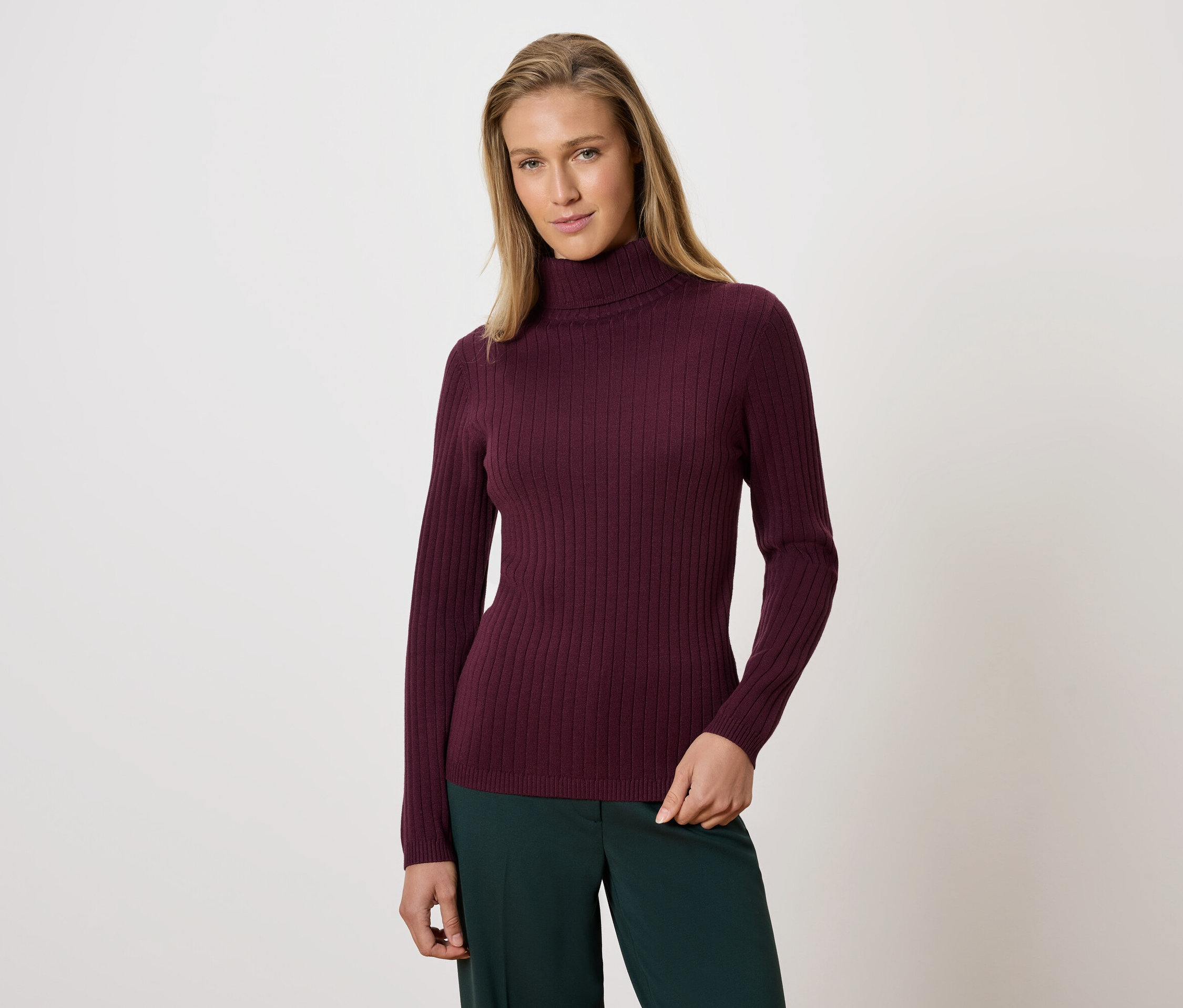 Femme posant portant un pull à col roulé bordeaux et un pantalon vert.