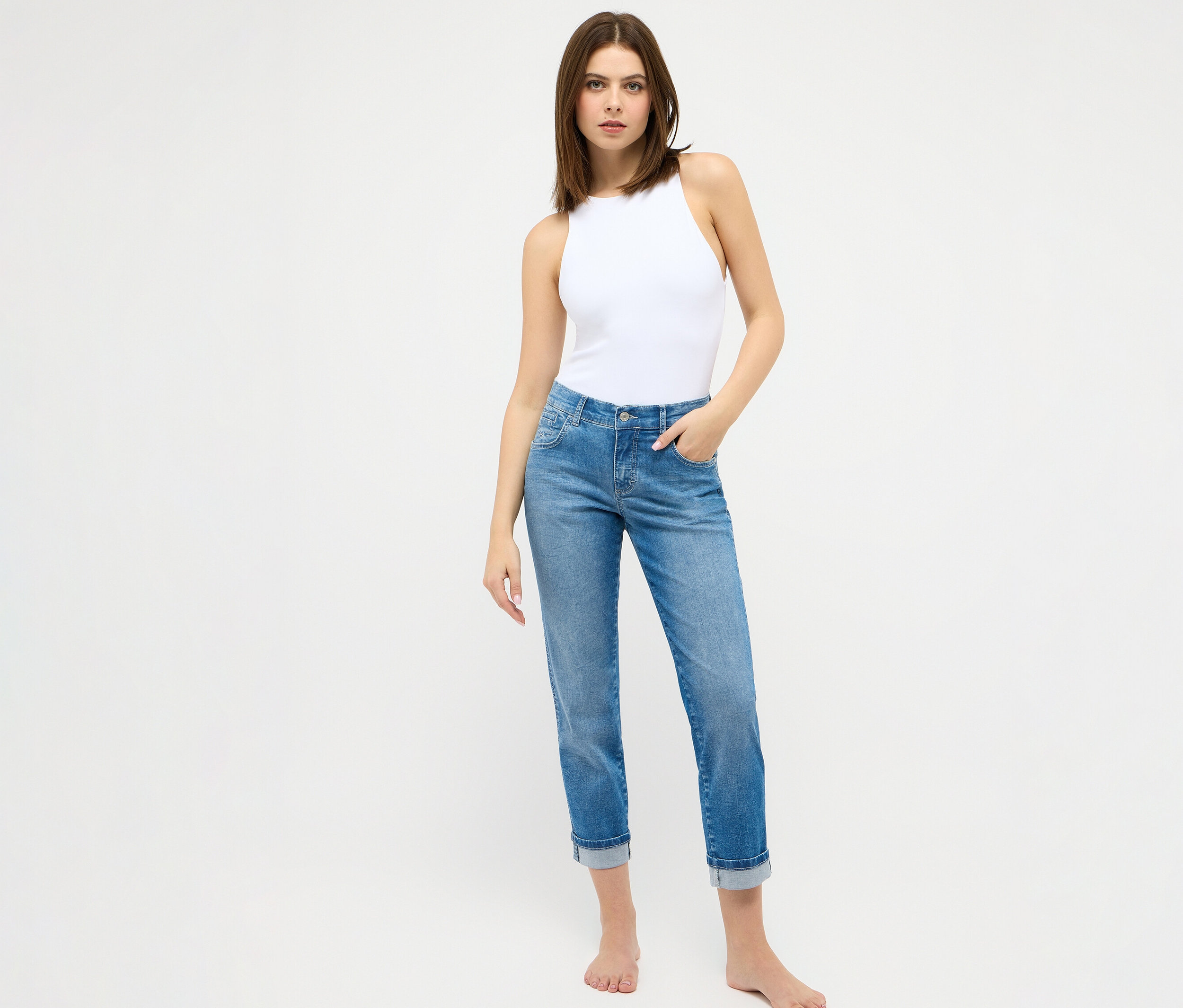 Femme debout pieds nus sur fond blanc portant un chemisier blanc et un jean Angels « Darleen » coupe cintrée 7/8 bleu clair.