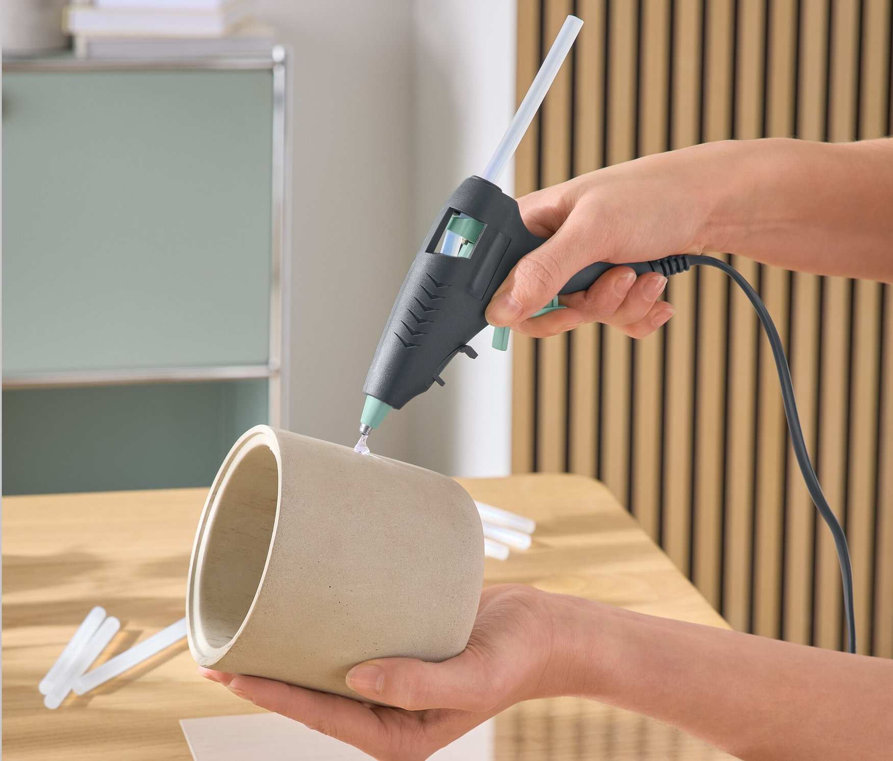 Une main utilise un pistolet à colle pour coller sur un pot de fleurs. Des coquillages sont sur la table.