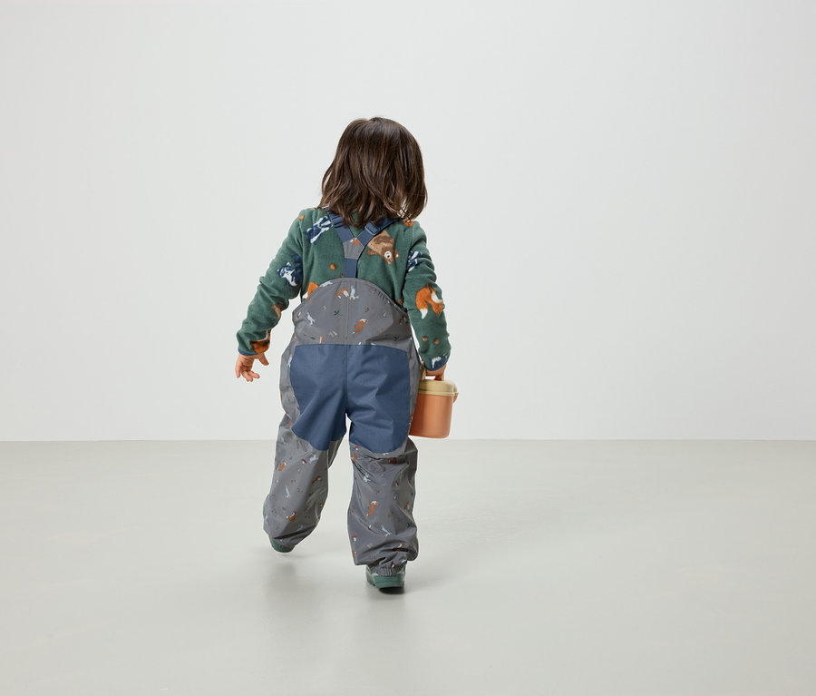 Un enfant, vu de dos, porte un pull vert à motif de renards et une salopette grise à imprimé, et tient un seau orange.