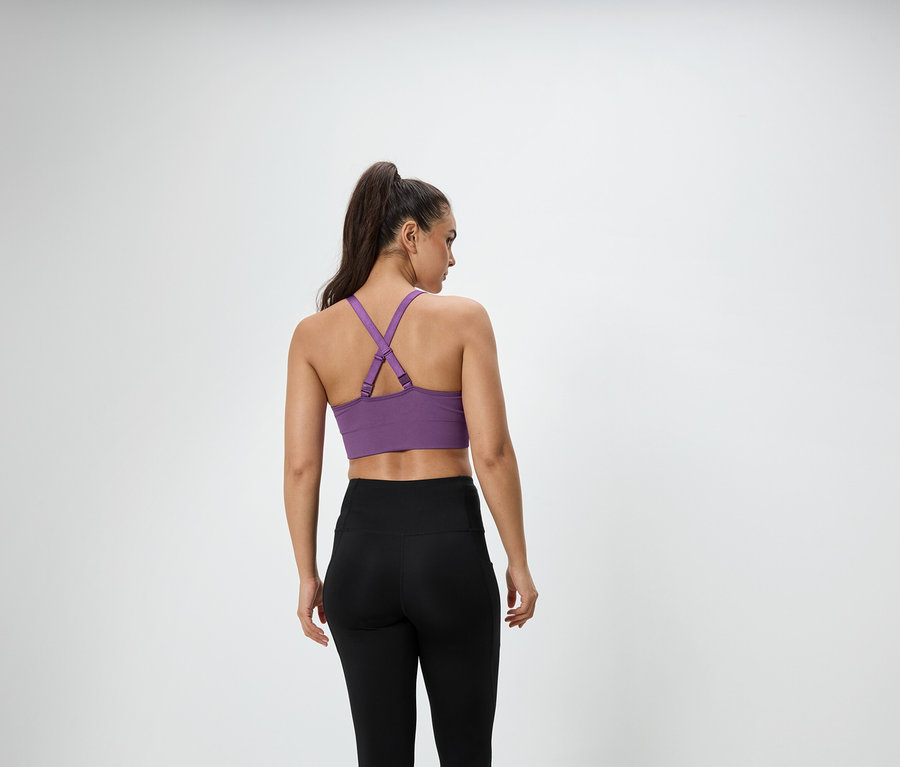 Une femme aux cheveux bruns attachés en queue de cheval se tient dos à la caméra, portant un soutien-gorge de sport violet et un legging noir.