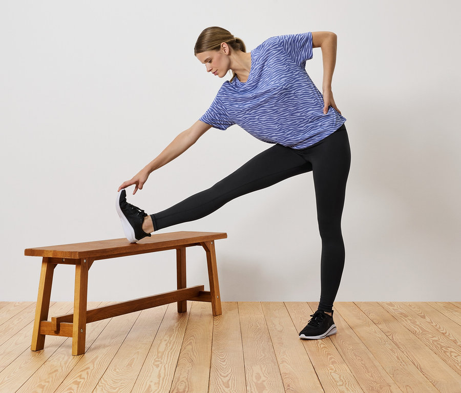 Une femme en t-shirt bleu et leggings noirs s'étire, une jambe posée sur un banc en bois.