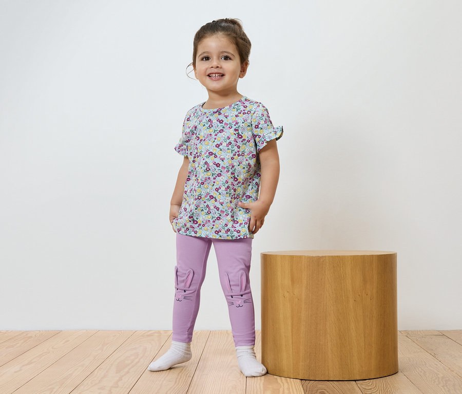 Une petite fille souriante se tient devant une table cylindrique marron, portant un t-shirt à fleurs et un legging violet.