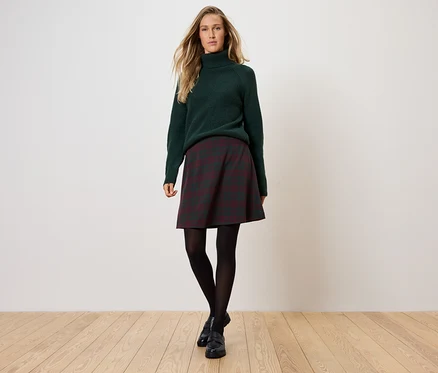 Une femme debout porte un pull à col roulé vert, une jupe à carreaux, des collants noirs et des chaussures noires.