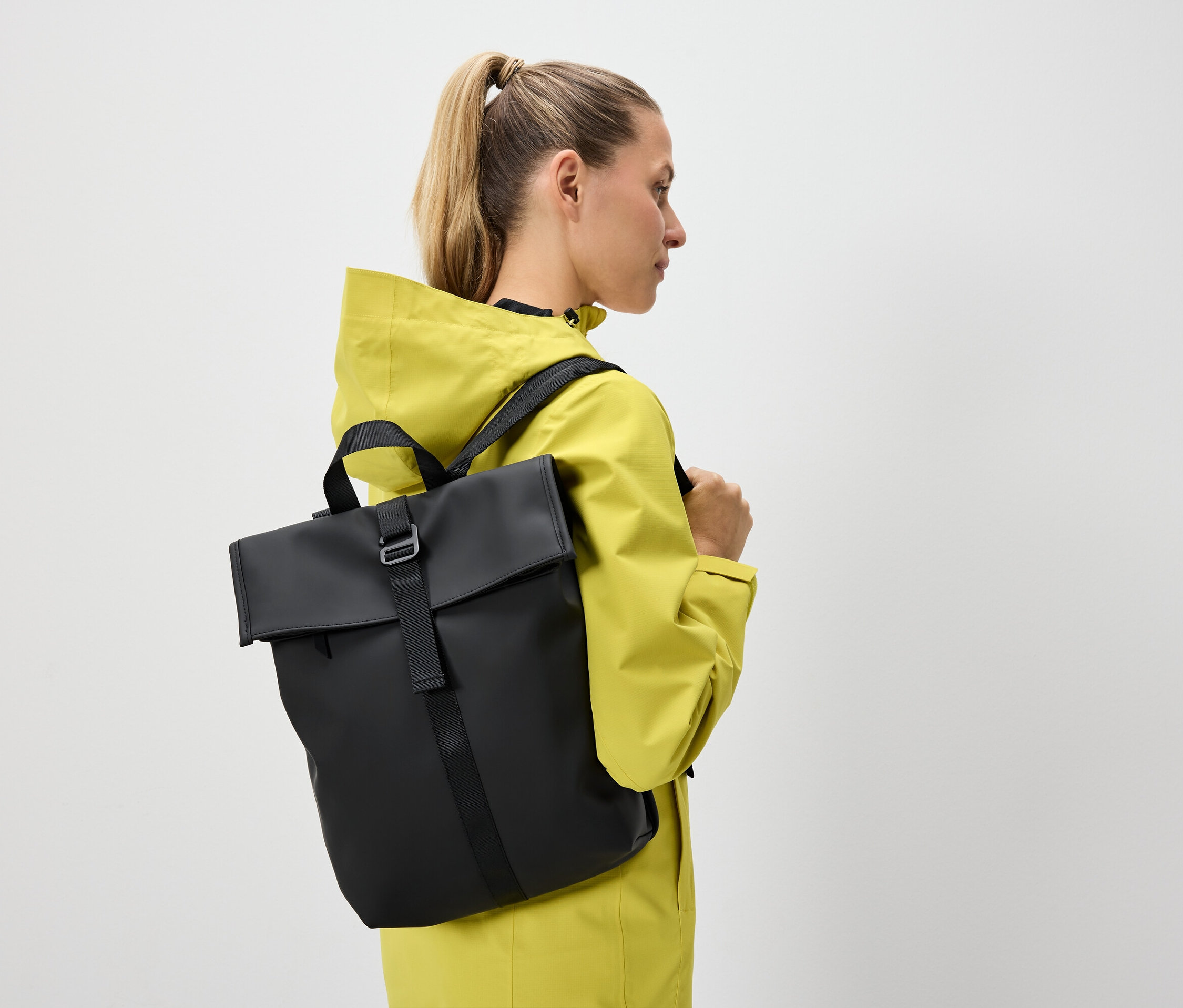 Une femme blonde aux cheveux attachés en queue de cheval porte une veste jaune et un sac à dos noir.