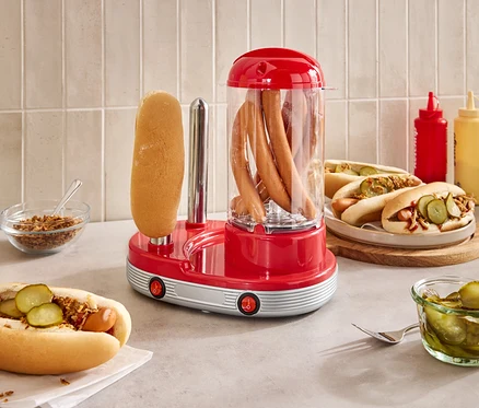 Sur un comptoir se trouve un appareil à hot-dogs rouge avec des saucisses à l'intérieur et un pain à l'extérieur.
