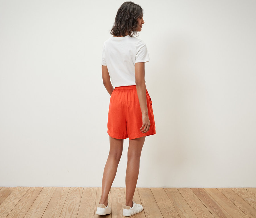 Une femme, vue de dos, porte un short en lin orange et un t-shirt blanc.