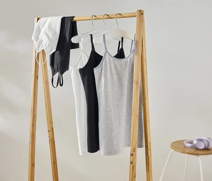 Un débardeur blanc, un débardeur noir, un caraco blanc, un caraco noir et un caraco gris sont suspendus à un portant en bois. Un casque violet est posé sur un tabouret à côté.