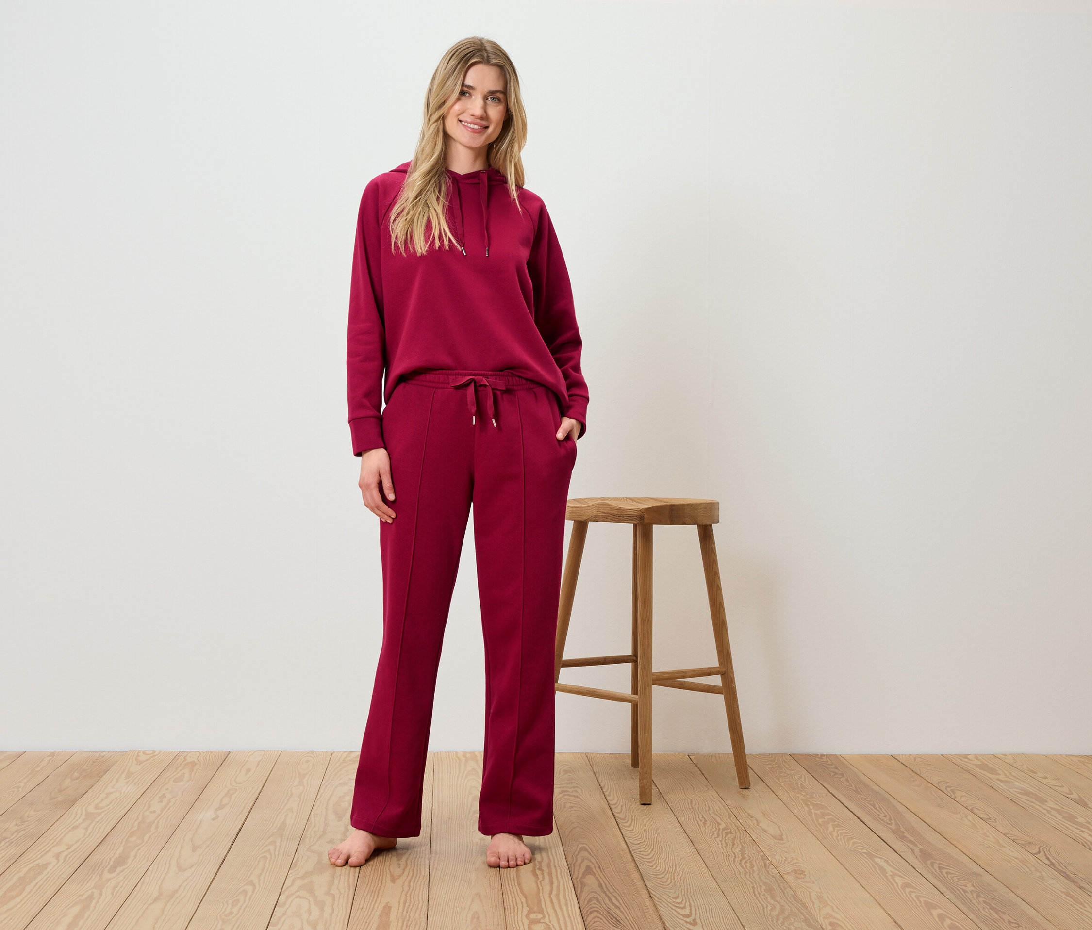 Une femme pose dans un ensemble bordeaux à capuche et pantalon devant un tabouret en bois.
