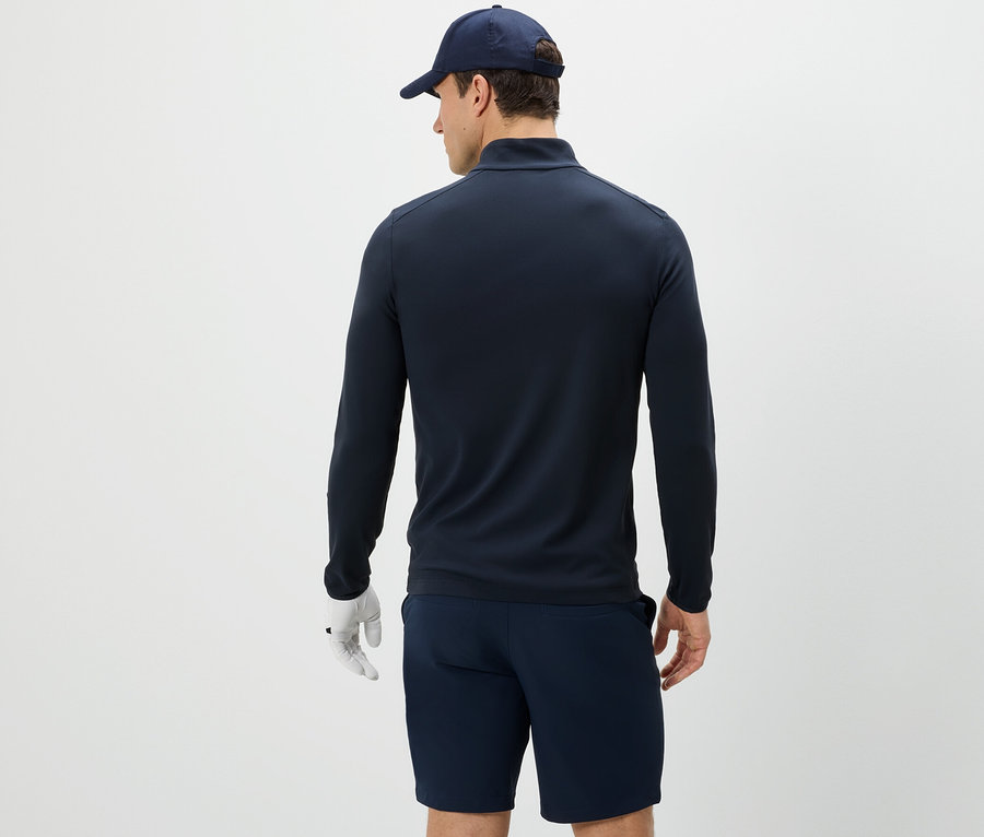 Un homme vu de dos porte une veste fonctionnelle, un short et une casquette de baseball.