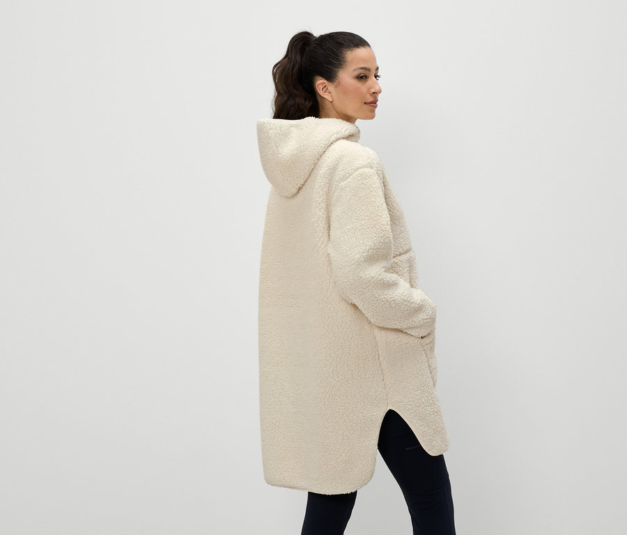 Une femme vue de dos porte un manteau à capuche couleur crème et a les mains dans les poches.