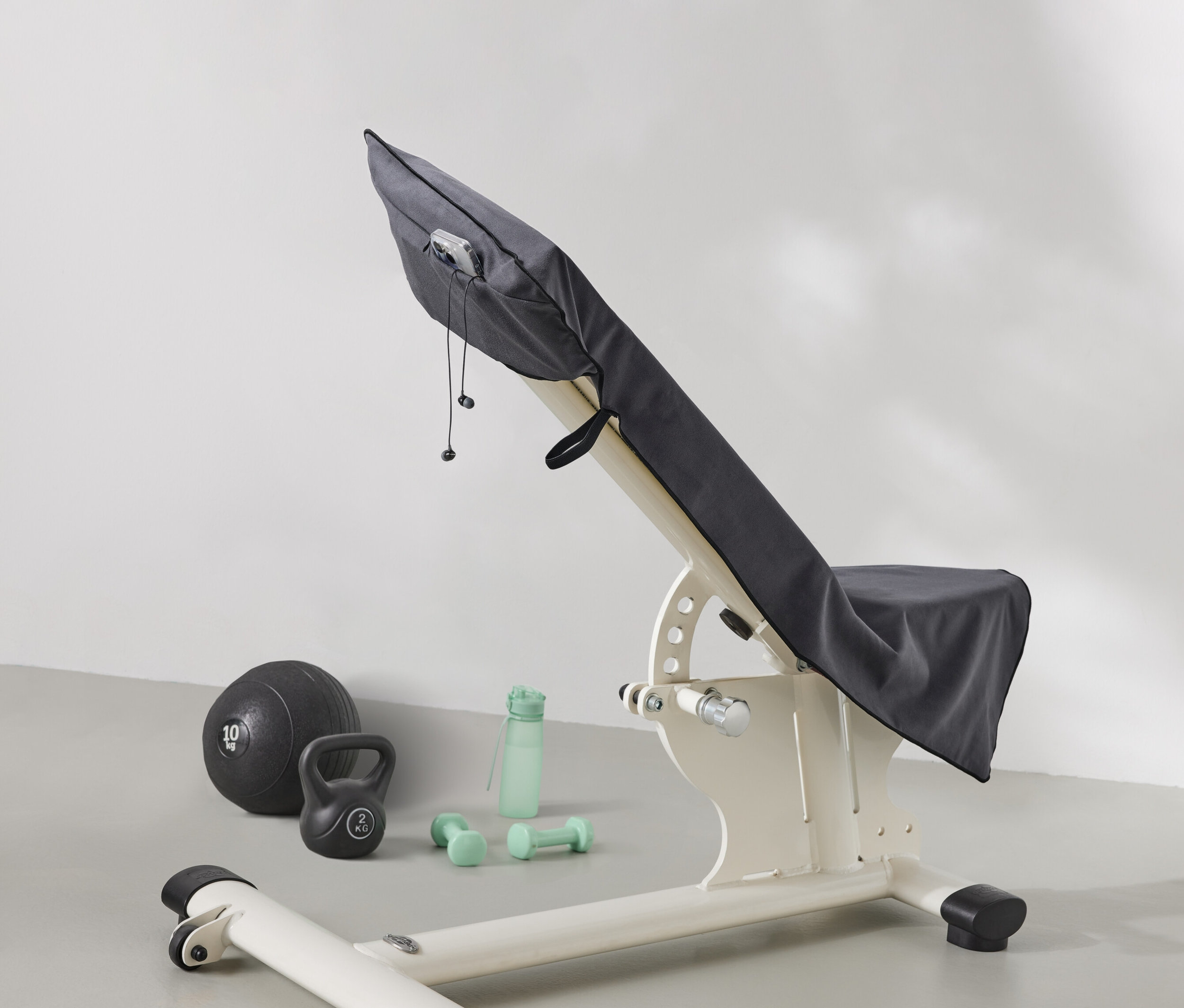 Un banc de musculation est recouvert d'un tissu gris. À côté du banc se trouvent un ballon de médecine, une kettlebell, une bouteille d'eau et des haltères.