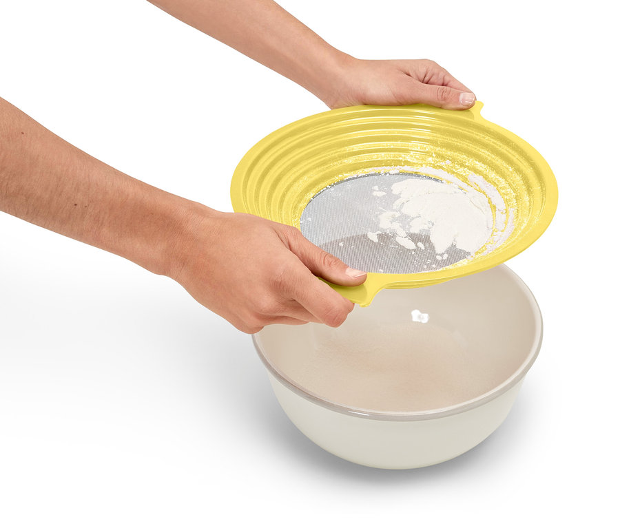 Une personne tient un tamis jaune au-dessus d'un bol et tamise de la farine.